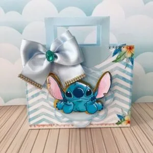 Arquivo Sacolinha Stitch Studio e pdf 28 Sacola de papel decorada com personagem azul, laço de cetim e flores tropicais