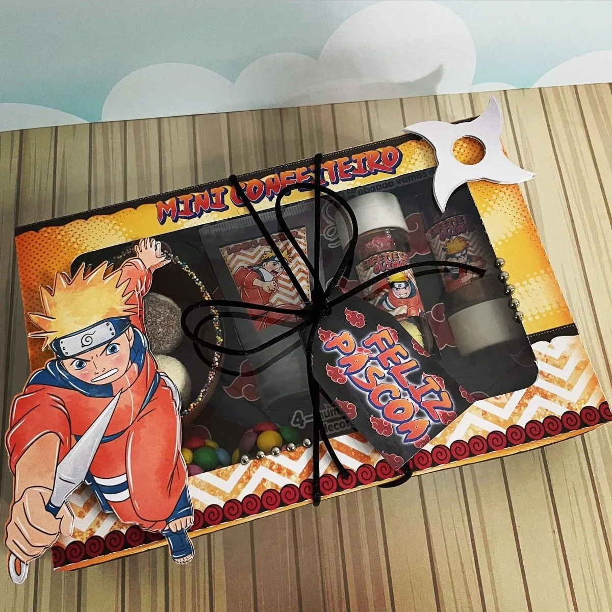 Arquivos De Corte Páscoa Naruto Mini Confeiteiro 6 Caixa decorada com personagem, doces, garrafinhas e laço preto sobre mesa.