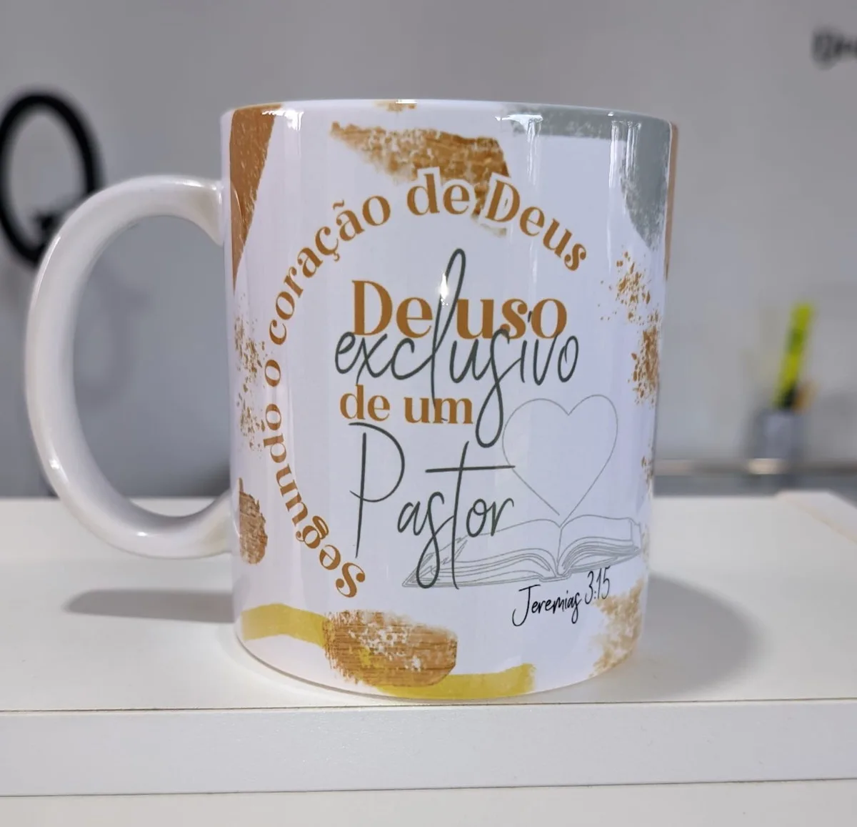 Caneca branca com estampa dourada contendo frases religiosas em fundo claro sobre mesa.