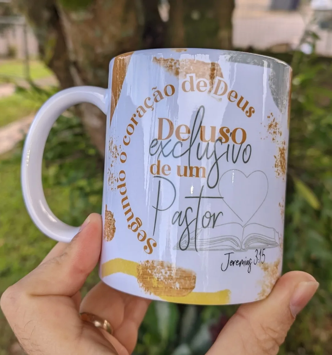 Mão segurando uma caneca branca com estampa religiosa, frases em português e detalhes dourados.