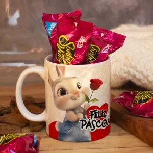 Caneca branca com coelho sorridente, rosa vermelha e doces embalados em papel vermelho.