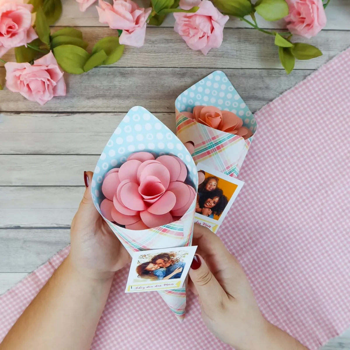 Mãos segurando cones de papel contendo flores artesanais de papel e fotos pequenas.
