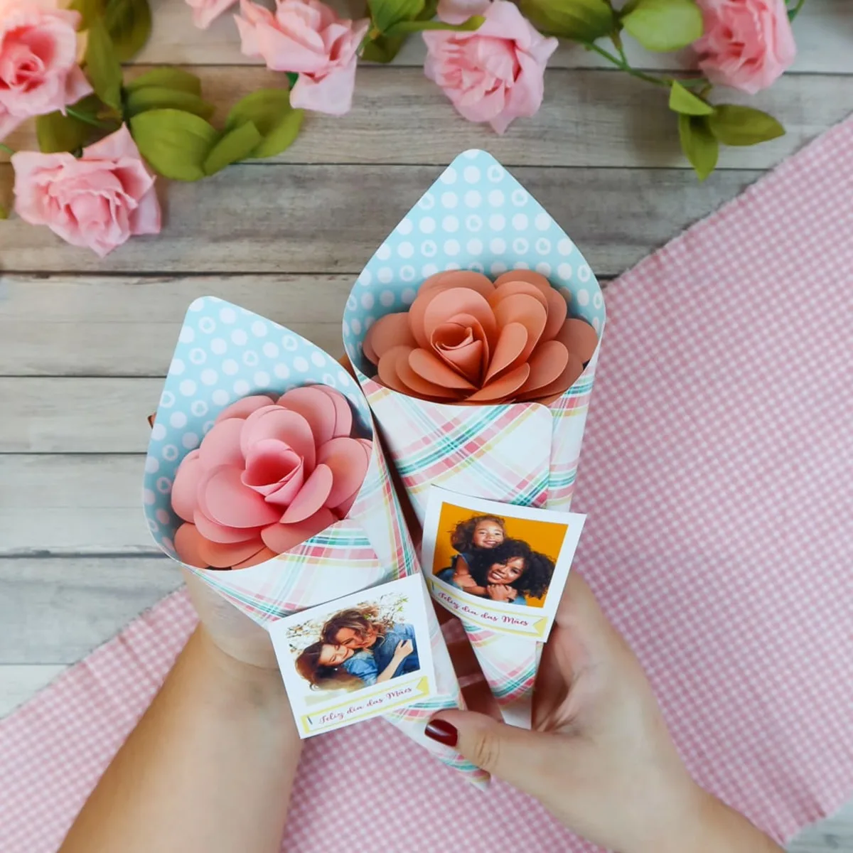 Mãos segurando dois cones de papel decorado contendo rosas artesanais e fotos pessoais.
