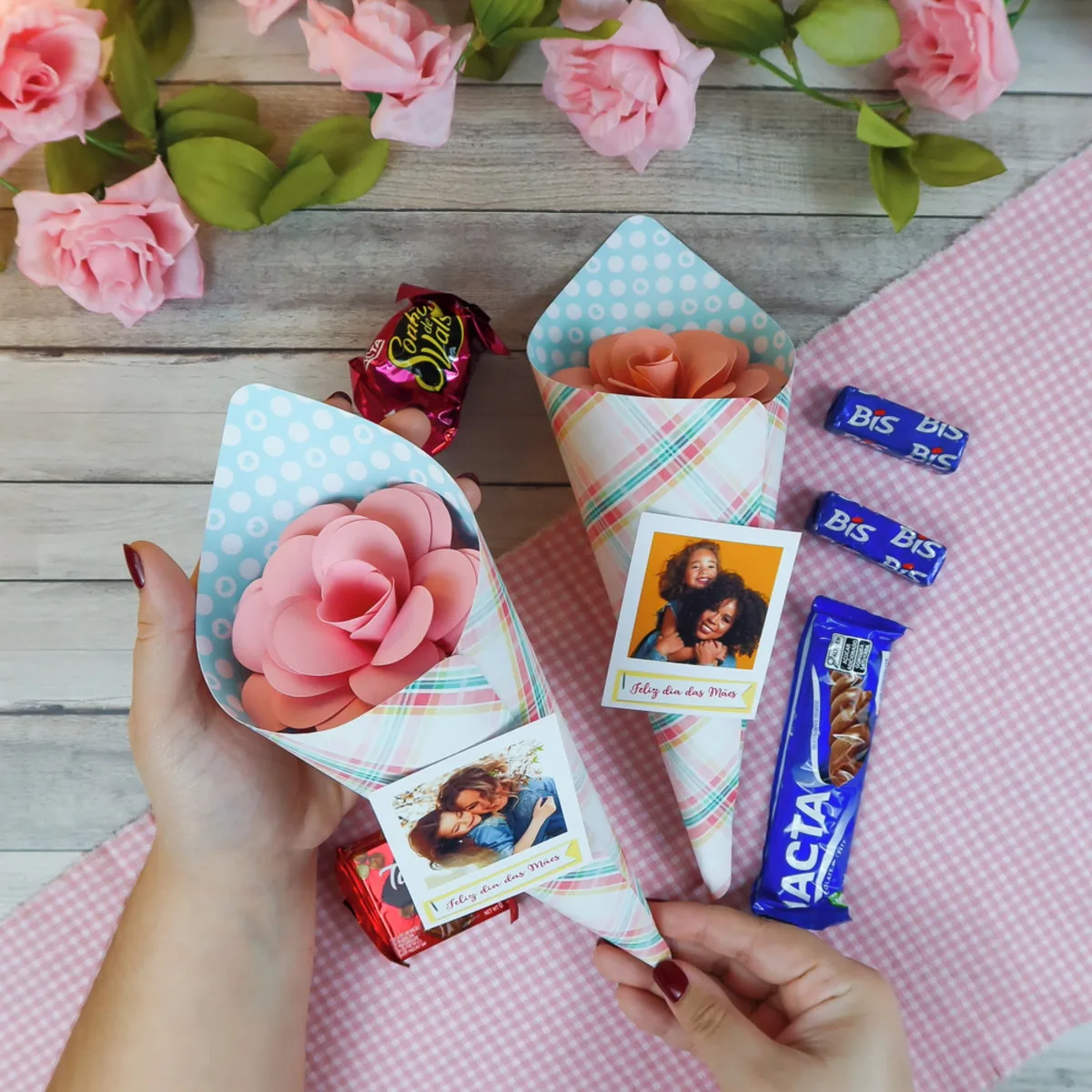 Mãos segurando cones de papel com rosas artificiais e fotos de dia das mães.