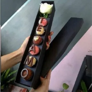 Caixa preta longa contendo doces confeitados e uma rosa branca.