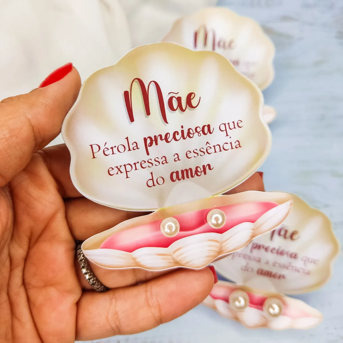 Mão segura cartões em formato de concha com a palavra Mãe e pérolas.