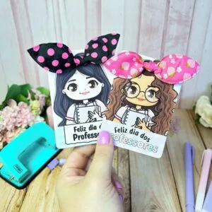 Card Xuxinha Dia dos Professores 18 Mão segura dois cartões ilustrados com personagens femininas e laços de tecido decorativos.