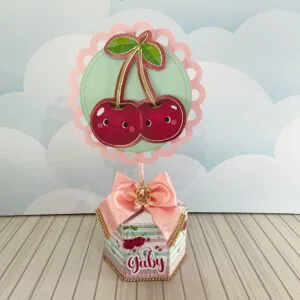 Decoração de mesa personalizada com tema de cerejas e laço rosa sobre caixa decorada.