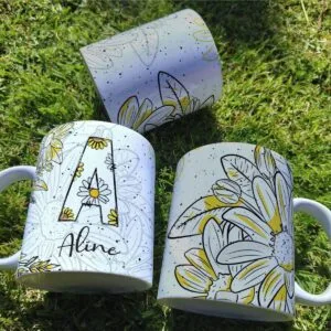 Três canecas brancas com estampa floral e letra A sobre gramado verde.