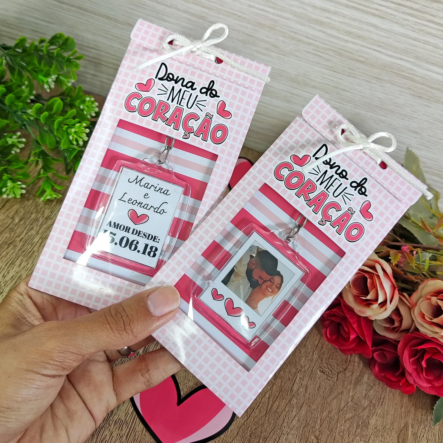 Mão segurando dois cartões personalizados com tema romântico, embalagens rosas e flores ao fundo