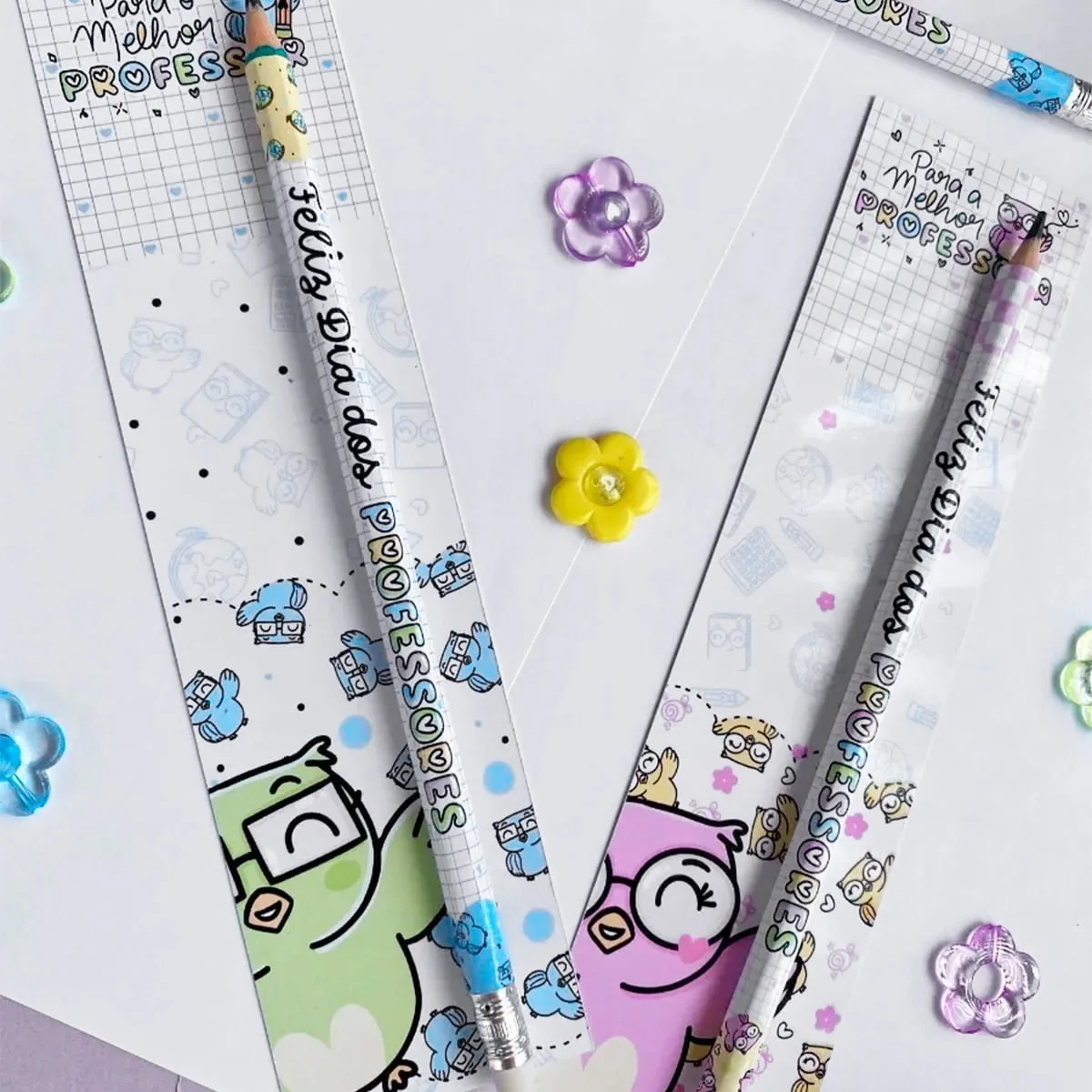Arte digital com dois lápis decorados, cartões e flores sobre fundo quadriculado.