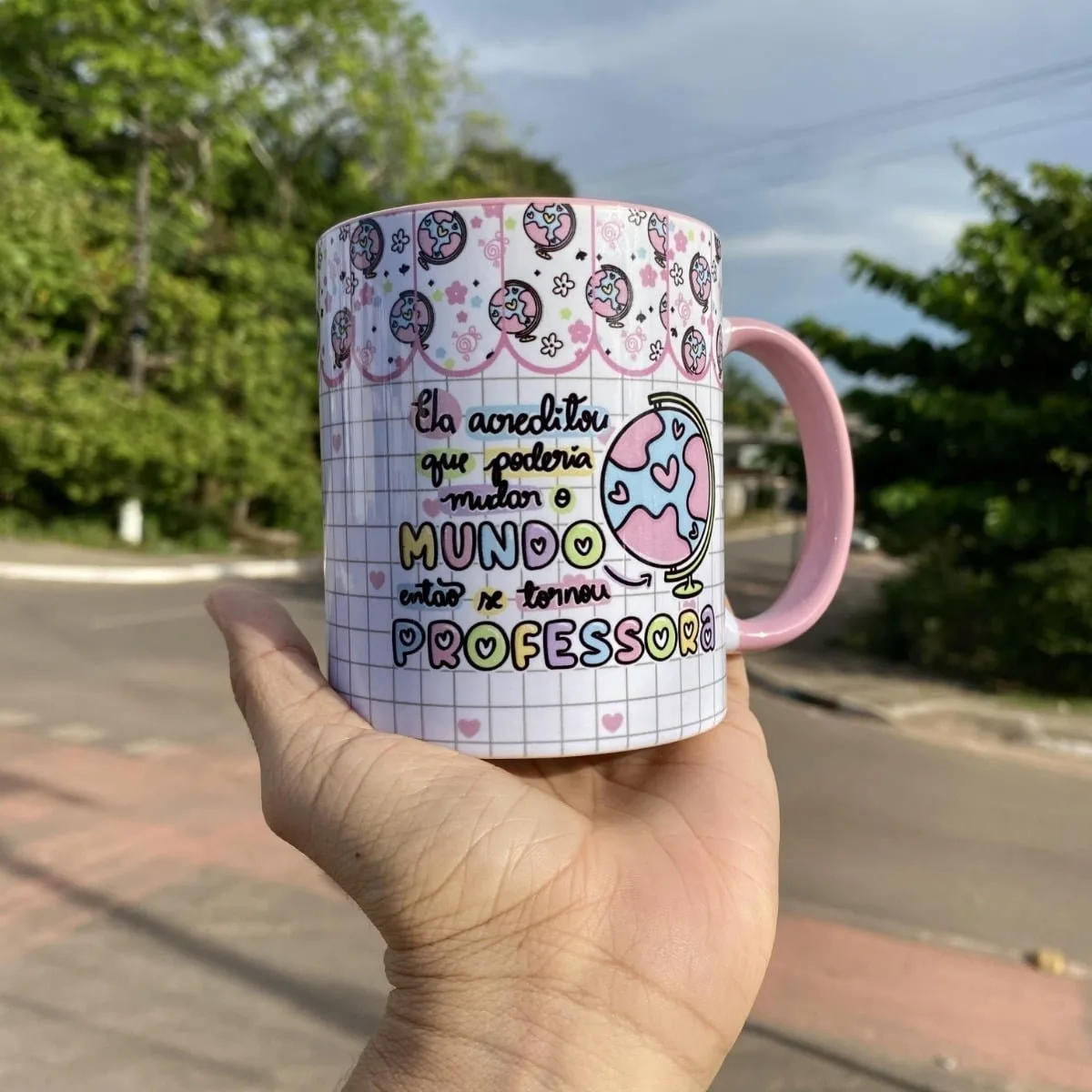 Mão segura caneca branca com frase sobre professora e globo, ao ar livre