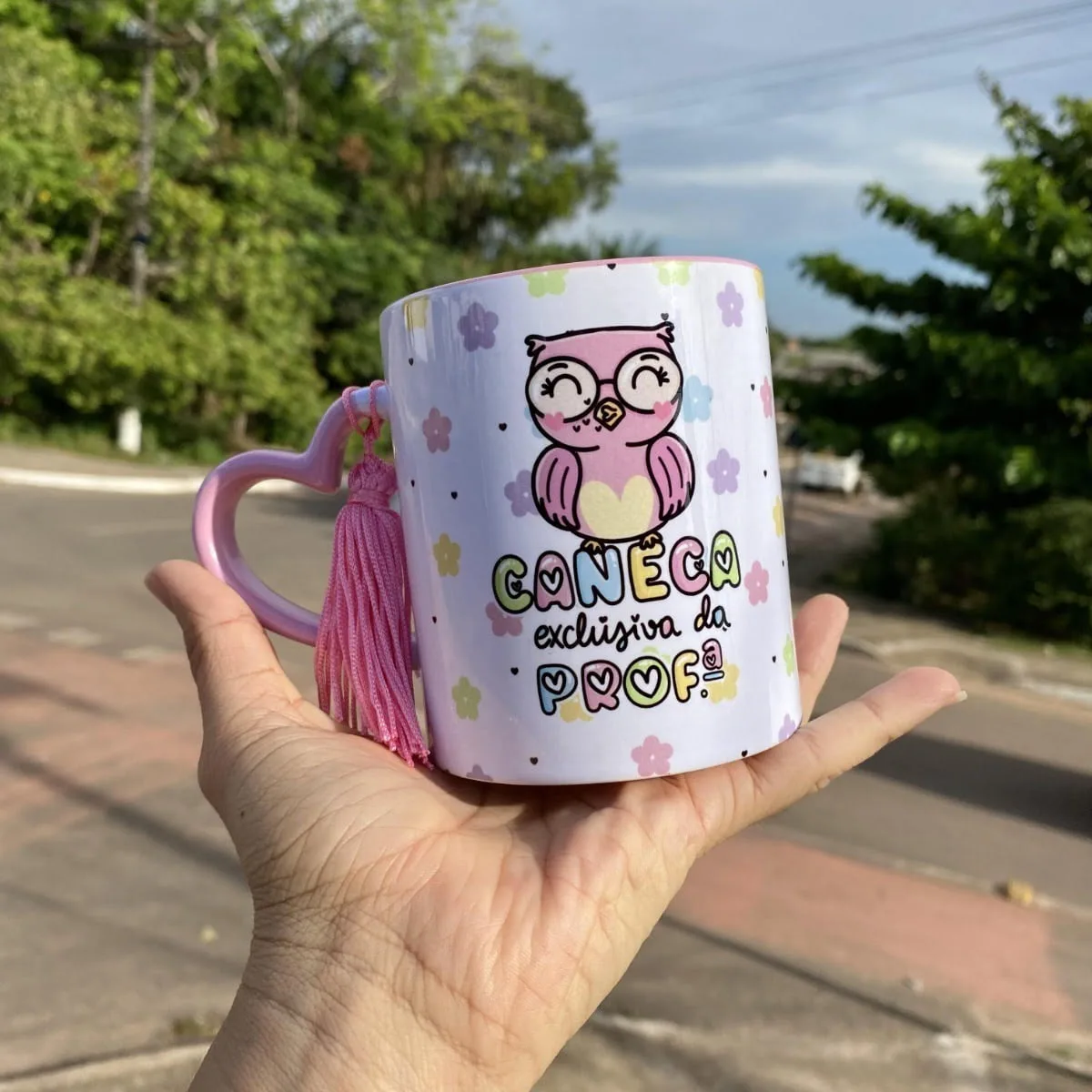 Mão segura caneca branca com coruja rosa e flores ao ar livre