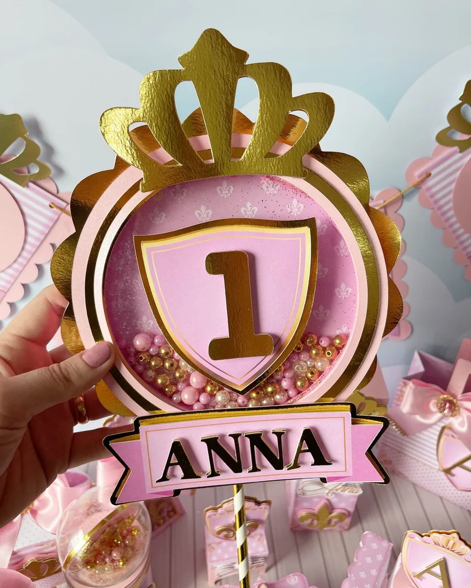 Kit Festa Realeza Menina 4 Mão segurando peça decorativa rosa e dourada com coroa, escudo e número um.