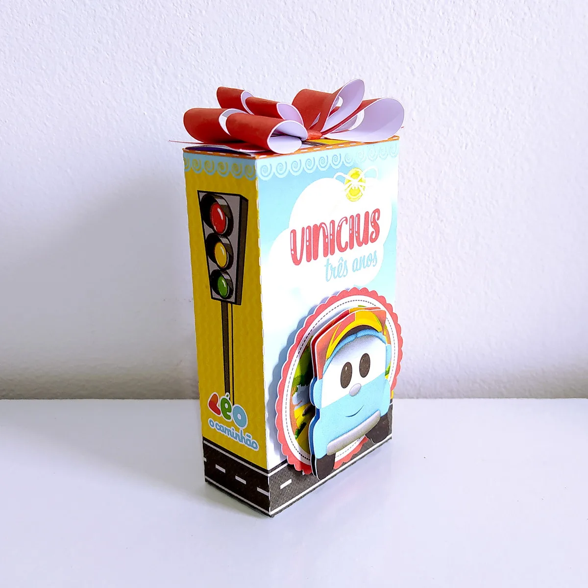 Kit Festa Só um bolinho Leo o Caminhão 3 Caixa de papel personalizada com tema de caminhão, semáforo e laço vermelho no topo.