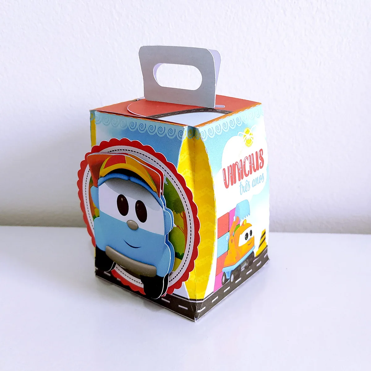 Kit Festa Só um bolinho Leo o Caminhão 4 Caixa de papel personalizada com ilustrações de carrinhos coloridos em um fundo branco.