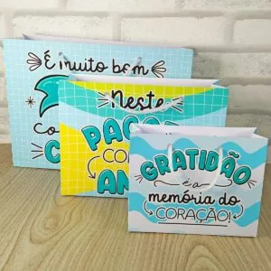 Tres sacolas de papel decoradas com frases coloridas em fundo azul e amarelo.