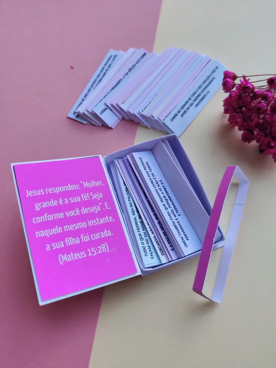 Caixa aberta cheia de pequenos papéis coloridos impressos com frases sobre fundo rosa.