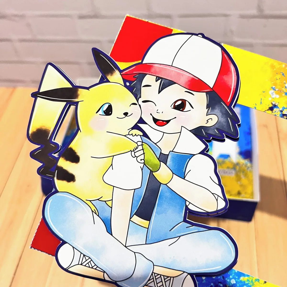 Mini Confeiteiro Páscoa Pokémon 3 Ilustração recortada em papel mostrando um garoto de boné abraçando seu ratinho amarelo.