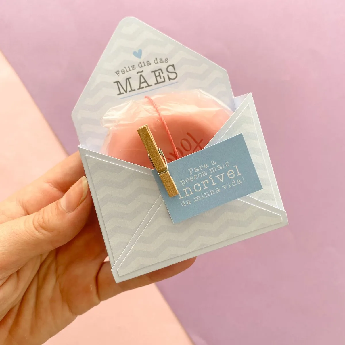 Mão segurando envelope de papel com cartão azul e sabonete rosa embrulhado