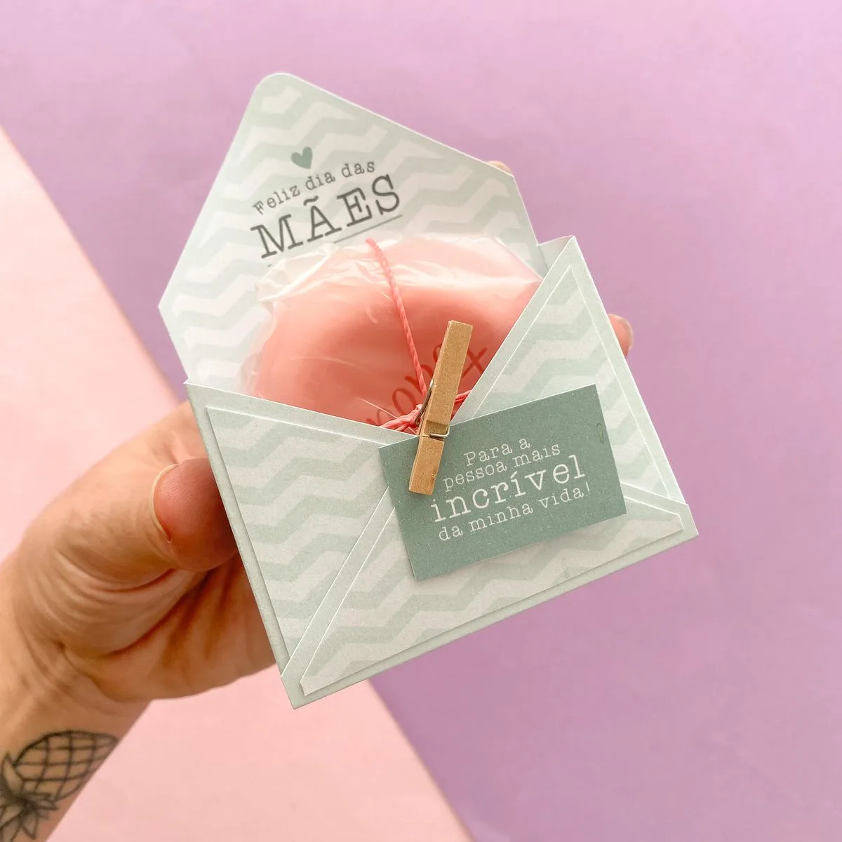 Embalagem de presente com objeto rosa embrulhado, tema Dia das Mães, segurada por uma mão.