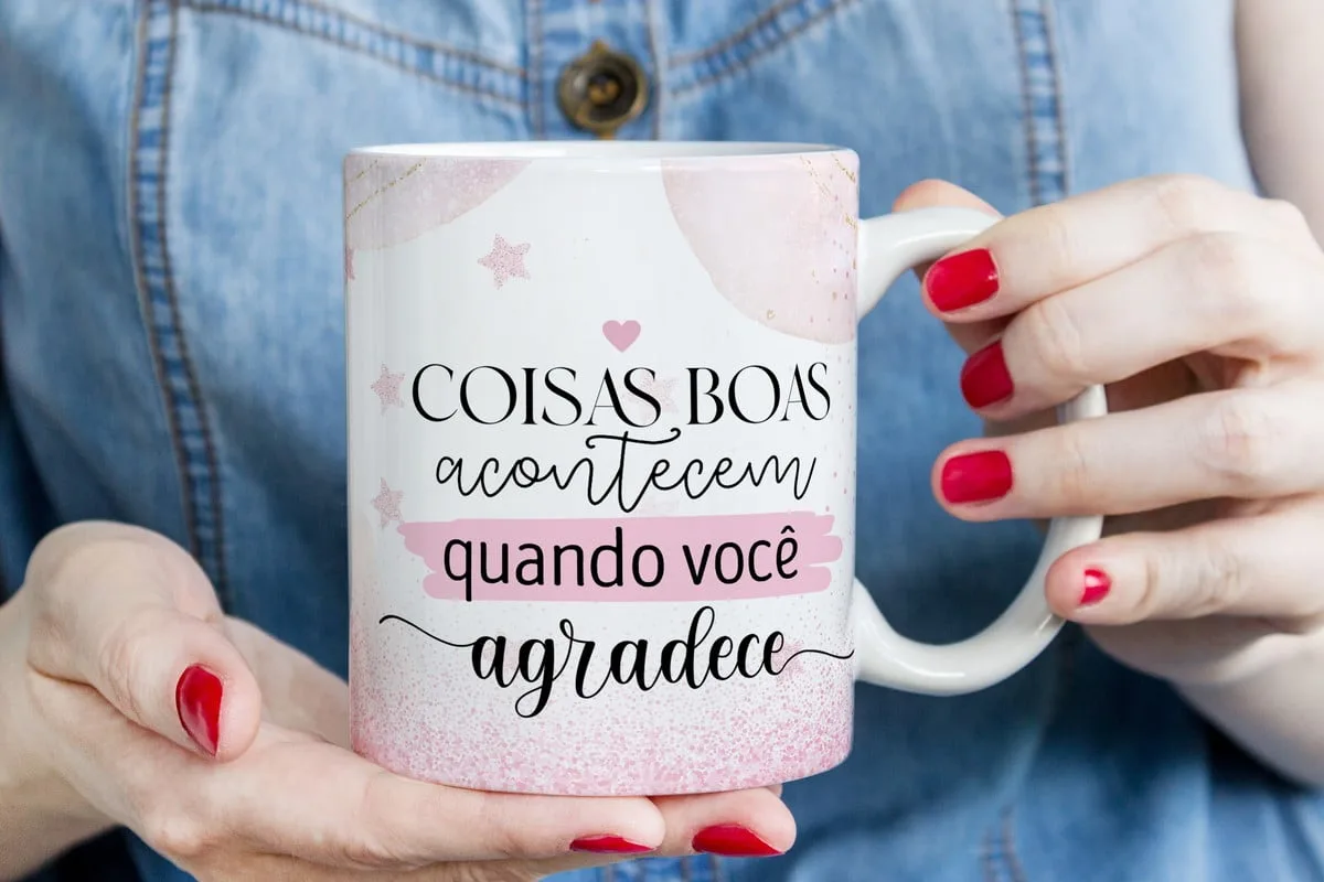 Pack Artes Canecas Gratidão 1 Caneca branca com frase motivacional e detalhes rosa, segurada por mãos com unhas vermelhas.