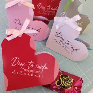 Porta Bombom Deus nos Seus Detalhes - pdf e Png 13 Caixinhas de papel em formato de coração com laços e doces sobre mesa.