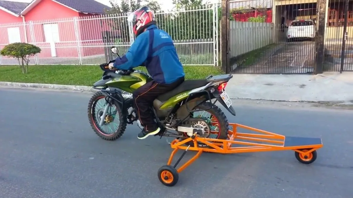 Motociclista em via asfaltada rebocando carreta laranja de estrutura metálica